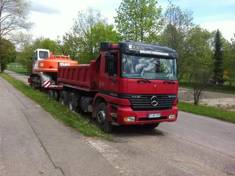 Roter Mercedes Kipper mit Tieflader und Bagger auf dem Weg zur Baustelle