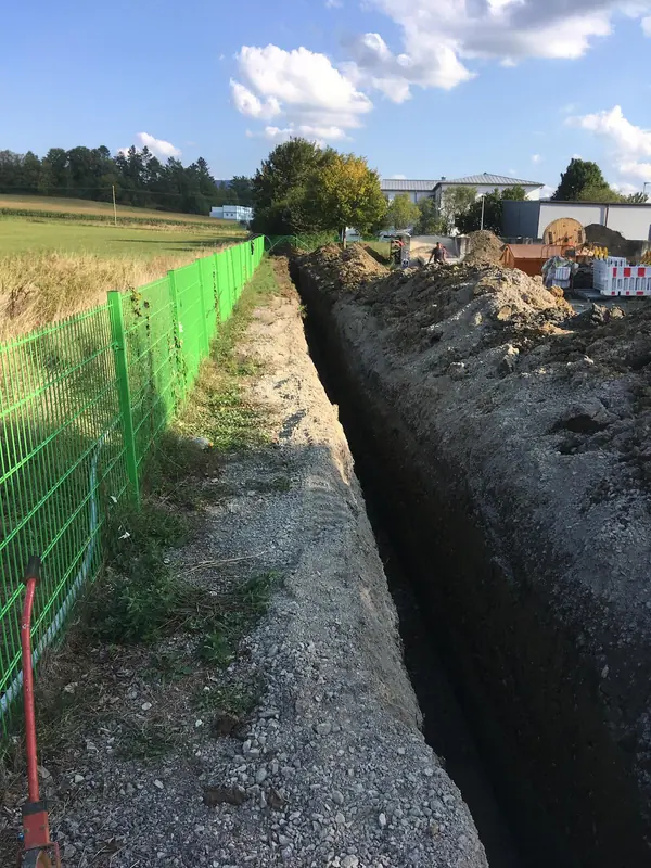 Tiefer, schmaler Kanalgraben entlang einer Grundstücksgrenze mit Bauzaun