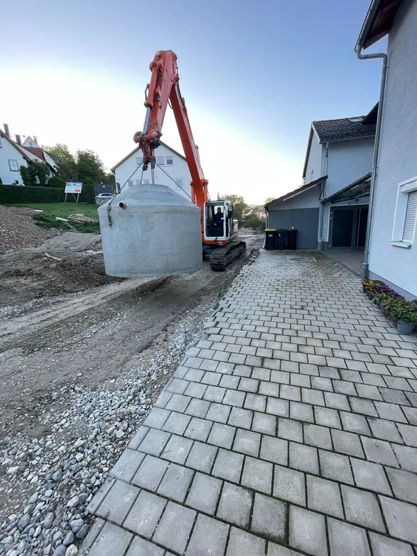 Bagger hebt einen schweren Betonschacht über einen fertigen Pflasterweg an die Einbaustelle