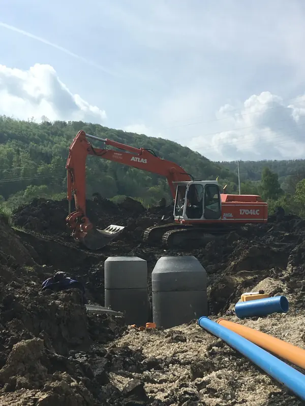 Atlas Bagger setzt große Behälter in die Baugrube, daneben liegen Kanal- und Wasserleitungsrohre