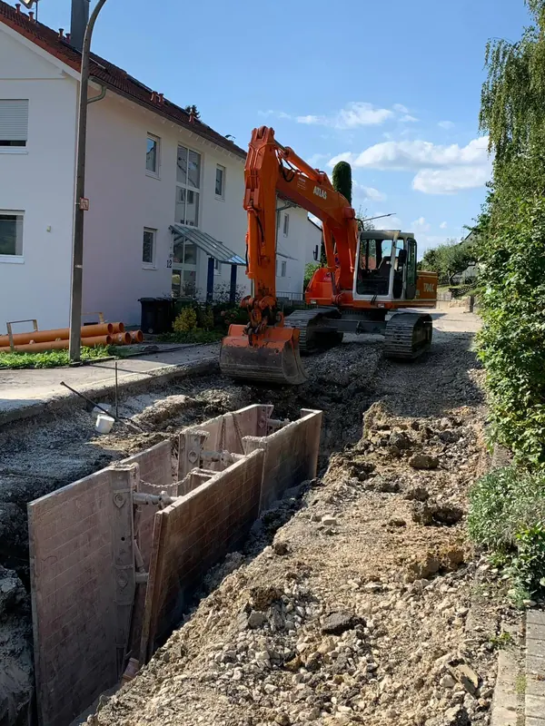Bagger arbeitet an einem mit Verbaubox gesicherten Graben zwischen Wohnhäusern